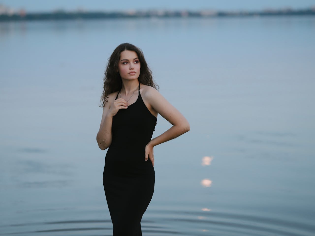 A stylish woman poses gracefully by the tranquil lakeside at twilight.