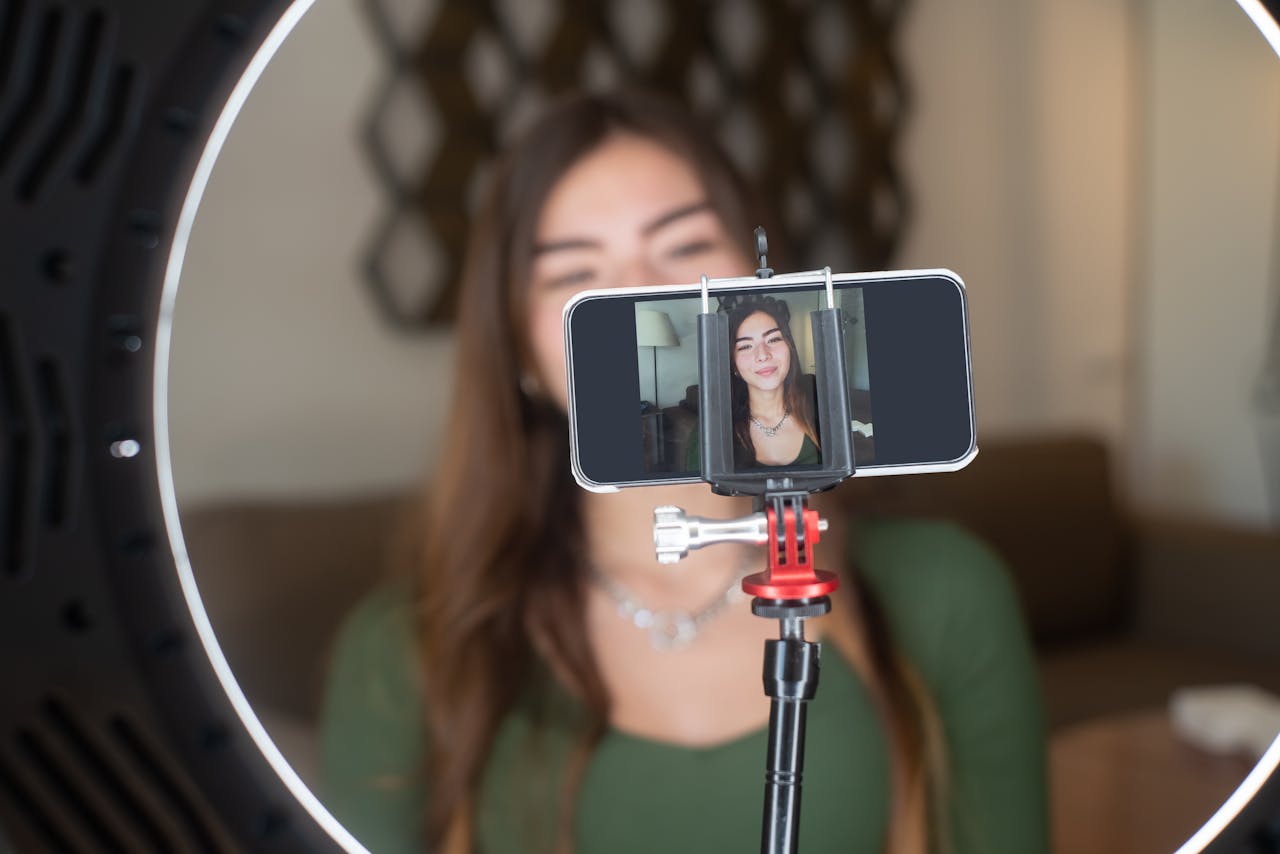 about-us A young woman live streams from home using a smartphone and ring light setup.